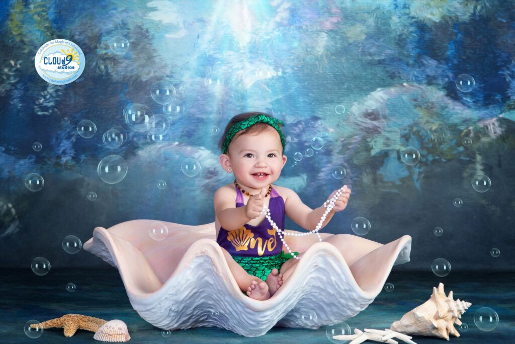 Baby girl in mermaid costume sitting in a sea-inspired photo set with ocean props at Cloud 9 Studios for her first birthday