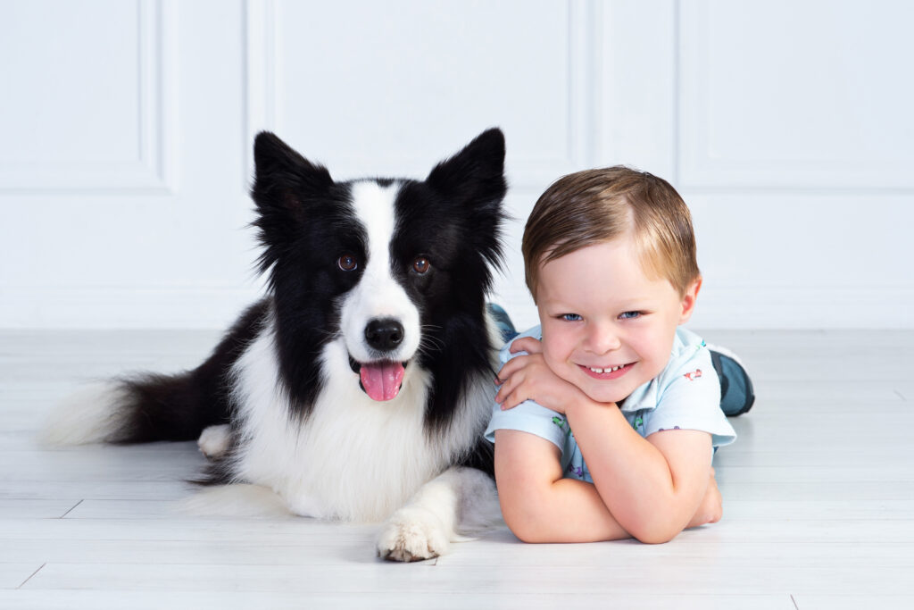 Young boy lying beside his dog in a studio portrait, both smiling at the camera during a child and pet photography session.