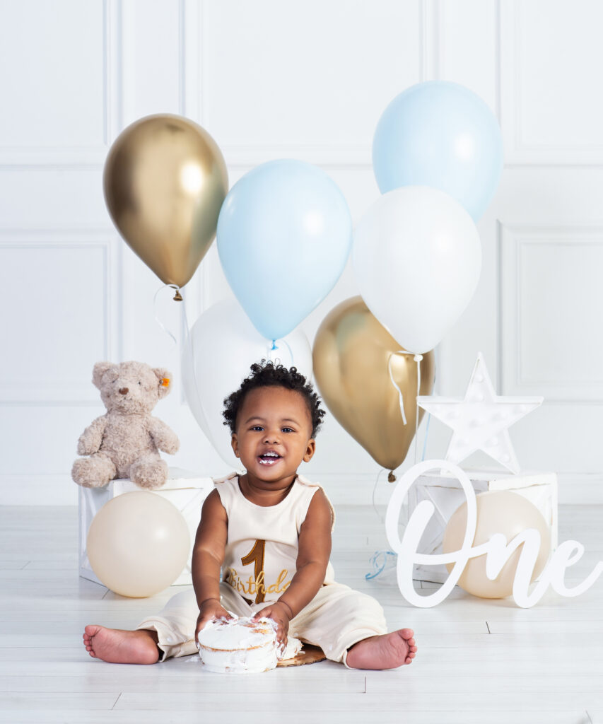 One-year-old baby smiling with blue and gold birthday balloons and a smash cake