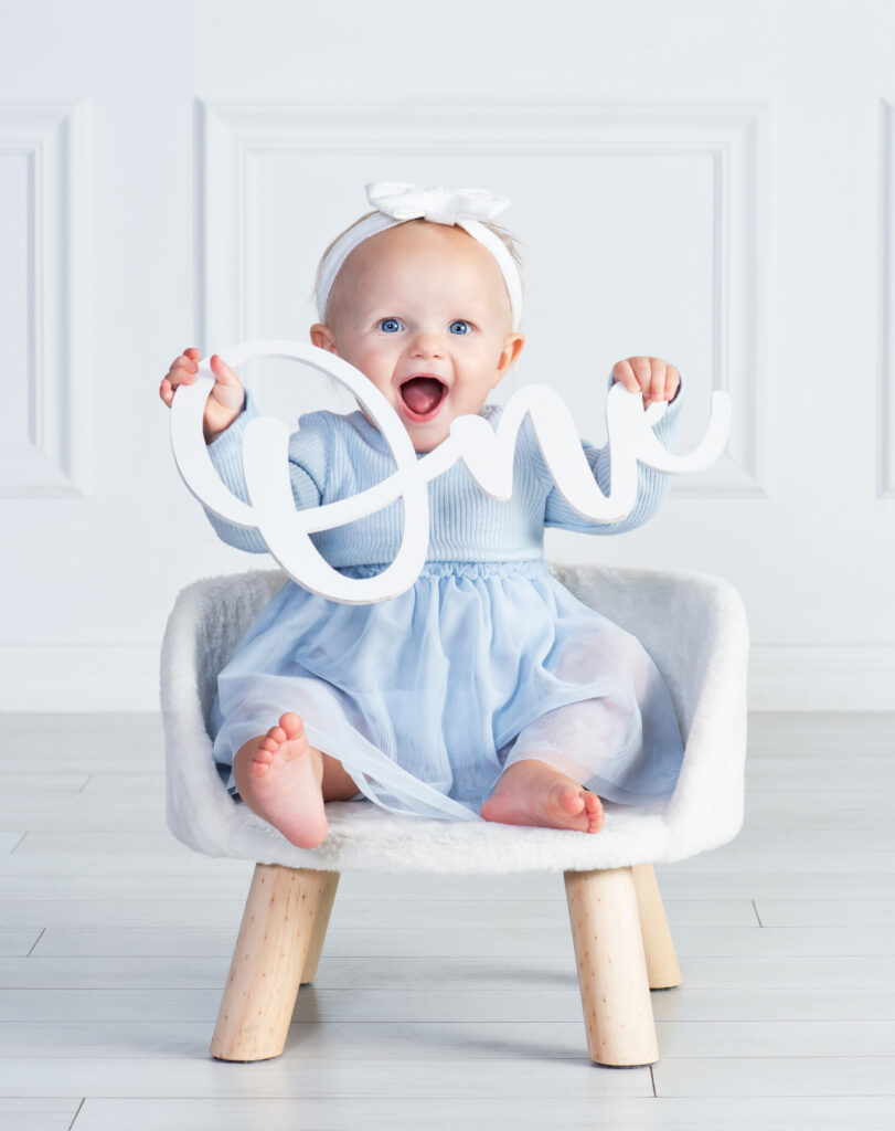 Smiling one-year-old baby in a simple classic portrait holding the word ONE at Cloud 9 Studios