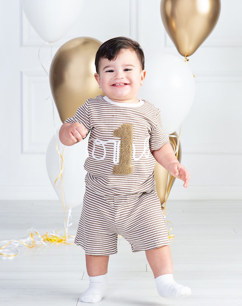Smiling one-year-old boy standing in a striped birthday outfit with a gold number one, surrounded by gold and white balloons.