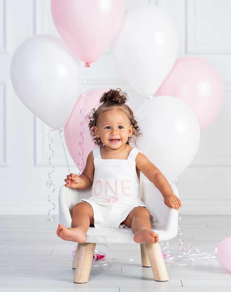 Smiling baby girl sitting on a small white chair in her first birthday outfit, surrounded by pink and white balloons.