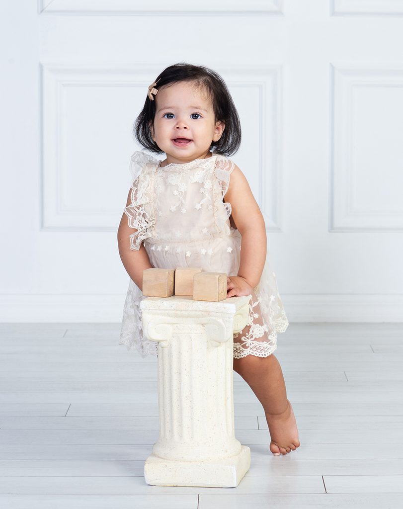 One-year-old girl in a cream lace dress standing while holding onto a decorative pedestal during her first birthday portrait session.
