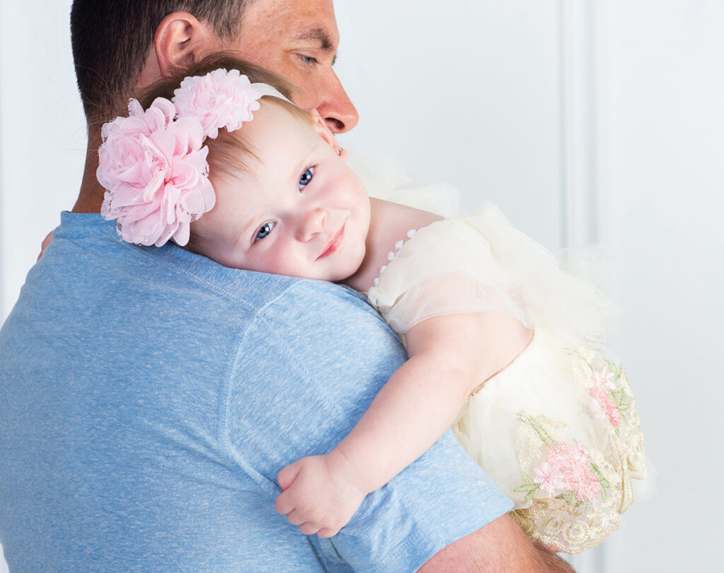 One-year-old girl snuggling on her dad’s shoulder during her first birthday portrait session