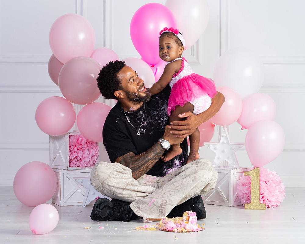 Dad covered in birthday cake frosting while holding his one-year-old daughter during her cake smash