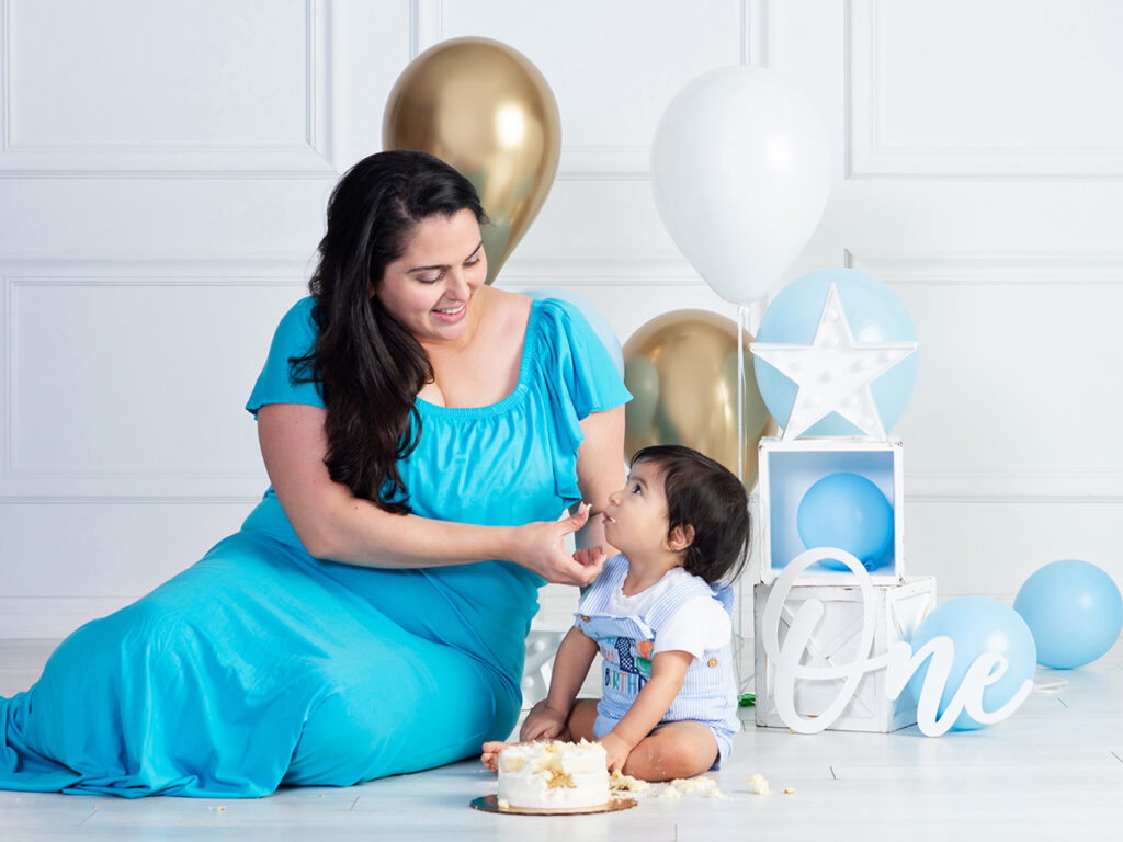 Mom sitting beside her one-year-old and helping him with his birthday cake during his portrait session