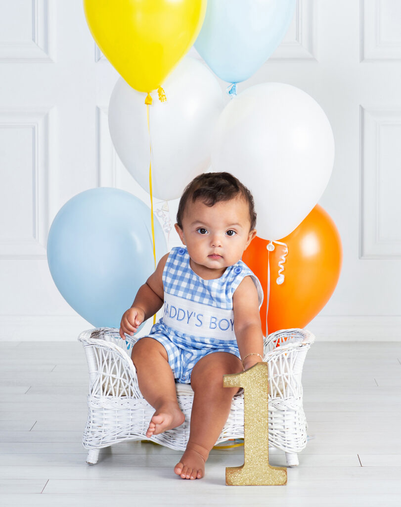 Serious one-year-old boy sitting with blue and orange balloons during his first birthday portrait session
